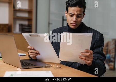 Concentrated mid aged brunette man entrepreneur in suit working in the office, sitting at the desk with laptop computer and documents, looking at pape Stock Photo