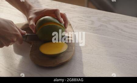 Man cutting ripe mango on olive board on white oak table, wide photo Stock Photo
