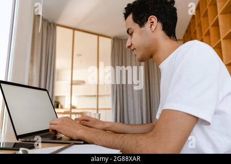 Pensive young hispanic man working studying on laptop computer while sitting at the desk at home Stock Photo