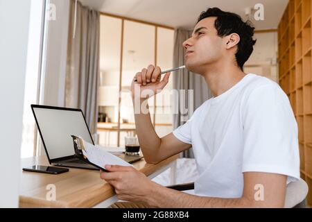 Pensive young hispanic man working studying on laptop computer while sitting at the desk at home Stock Photo