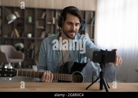 Happy young man in headphones recording video playing on guitar. Stock Photo