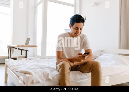 Smiling young hispanic man using mobile phone while sitting on bed at home Stock Photo