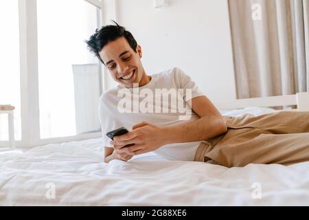 Smiling young hispanic man using mobile phone while laying on bed at home Stock Photo