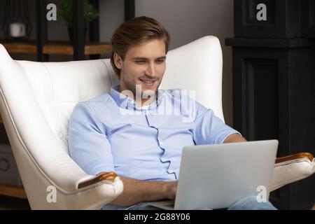 Young bearded man sit in large armchair work on laptop Stock Photo