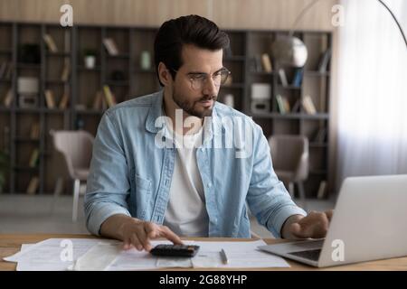 Concentrated young man calculating domestic expenditure, paying bills online. Stock Photo