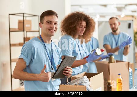 Portrait of happy male volunteer smiling at camera, showing thumbs up and holding tablet pc while sorting, packing food stuff in cardboard box Stock Photo