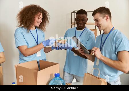 Team of diverse young volunteers in protective gloves smiling while sorting, packing foodstuff in cardboard box, working together on donation project Stock Photo
