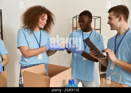 Team of diverse young volunteers in protective gloves smiling while sorting, packing foodstuff in cardboard box, working together on donation project Stock Photo