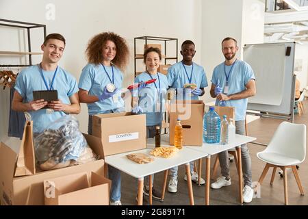 Full length shot of happy volunteers smiling at camera while sorting, packing foodstuff in cardboard boxes, working together on donation project Stock Photo