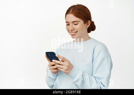 Happy girl chatting with friend on mobile phone. Cute young redhead woman using smartphone app, smiling at cellphone screen, shopping online, standing Stock Photo