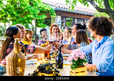 Happy multi ethnic friends having fun outdoor toasting red wine glass at winery on sunset - Young people enjoying time together at table in the restau Stock Photo