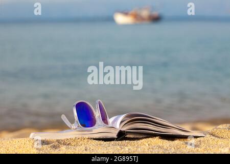 Open book and sun glasses on the beach with small ship in the background. Concept for a relaxing, sunny day near the sea.. Stock Photo