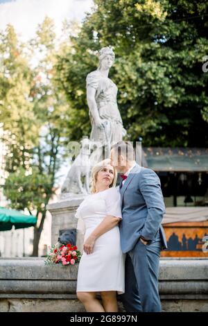Front view of a mature couple hugging in front of a water fountain, looking each other with love and dreaming. Pretty blond woman holding a bouquet of fresh flowers. City square view Stock Photo
