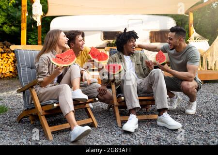 Joyful diverse friends chatting near camper van, eating yummy watermelon, sitting in lounge chairs, laughing outdoors Stock Photo