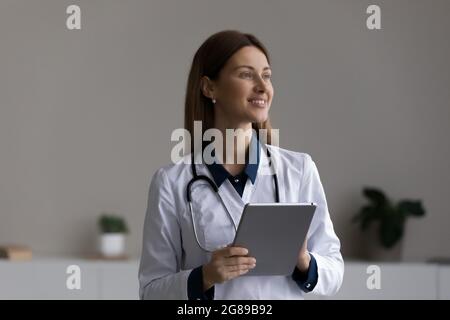 Happy thoughtful female doctor, physician using tablet computer at office Stock Photo