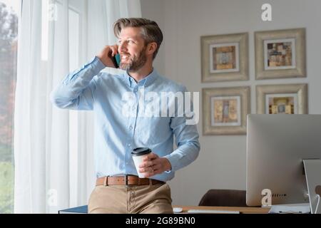 Handsome businessman having a business call while standing at office desk while on coffee break. Stock Photo