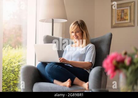 Blond haired smiling woman using her laptop while sitting at the armchair and working from home. Home office. Stock Photo