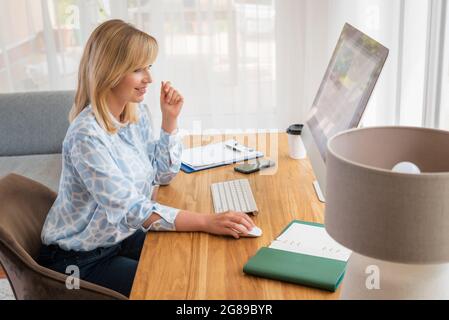 Smiling blond haired businesswoman sitting at desk and using computer while working from home. Home office. Stock Photo