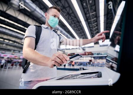 Man using self service check-in machine. Passenger scanning ticket on smart phone at airport terminal. Selective focus on hand holding smartphone. Stock Photo