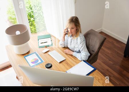 Shot of blond haired businesswoman sitting at desk and having a phone call while working from home. Home office. Stock Photo