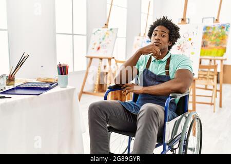 Young african american artist man sitting on wheelchair at art studio looking confident at the camera with smile with crossed arms and hand raised on Stock Photo