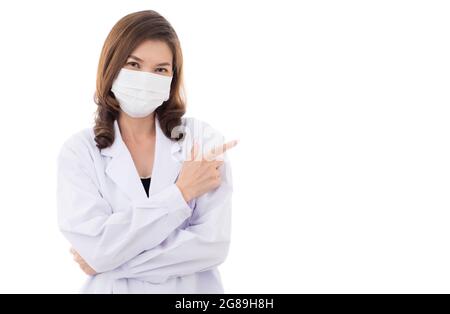 Pretty beautiful Asian woman in doctor gown or lab coat wearing surgical hygiene mask standing and pointing finger in advertising pose to introduce so Stock Photo