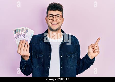 Young hispanic man holding egyptian pounds banknotes smiling happy pointing with hand and finger to the side Stock Photo