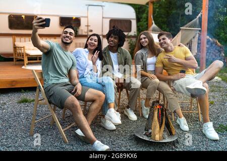 Diverse young friends taking selfie near campfire in evening, using smartphone, sitting next to RV during camping trip Stock Photo