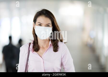 Beautiful adult middle age Asian working office businesswoman wearing surgical protective medical mask on face looking straight to camera while walkin Stock Photo