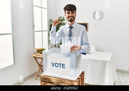 Hispanic man with beard voting putting envelop in ballot box smiling and confident gesturing with hand doing small size sign with fingers looking and Stock Photo