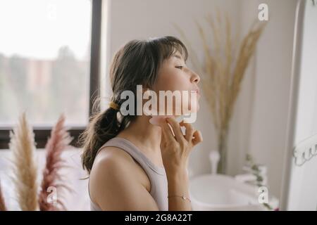 Beautiful asian woman doing face massage with quartz scraper during daily beauty morning routine Stock Photo