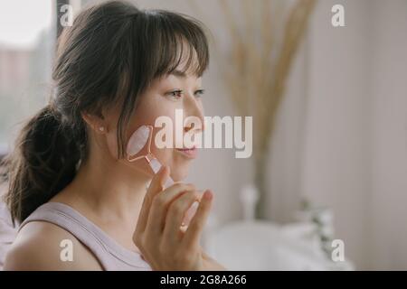 Asian woman using pink roller Gua Sha facial massage. Anti age, face yoga, lifting concept Stock Photo