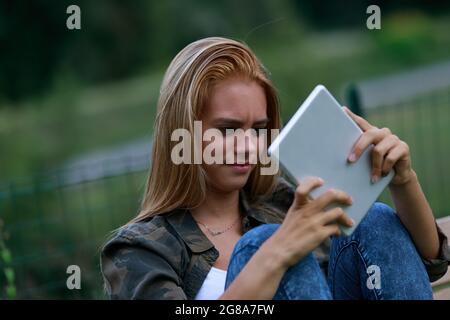 Confused or sceptical young woman staring at her handheld tablet with a perplexed frown as she sits relaxing outdoors in evening light Stock Photo