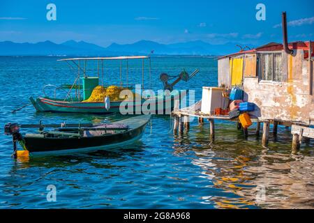 Sunlit small blue, white and yellow Mediterranean fishing boats, with sparkling turquoise sea and pale blue sky. Hut by the seaside. Stock Photo