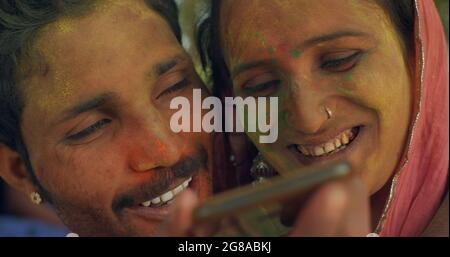 A cheerful Indian couple is sending a voice message over the phone; a man and woman with painted faces are attending the Holi festival Stock Photo