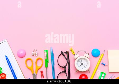 School notebook and office supplies on a pink background. Back to school abstract background. White alarm clock and multi-colored pens and black glasses. Stock Photo