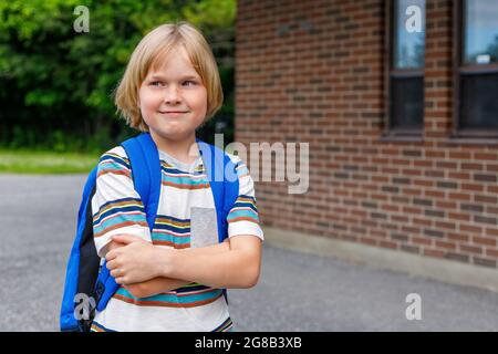 Beautiful happy smiling child near school building at the schoolyard. Little blond student carrying blue backpack. Back to school concept. Stock Photo