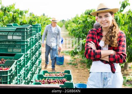 Smiling young woman farmer standing in orchard during harvest of sweet cherries Stock Photo