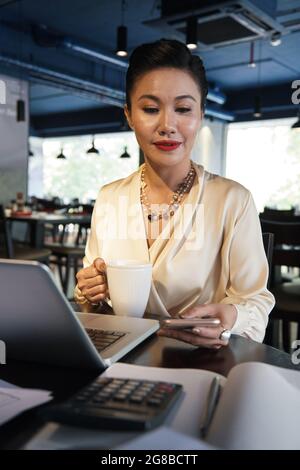 Business lady in silk blouse working on laptop at restaurant table and checking notifications or answering text messages on her phone Stock Photo