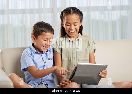Happy brother and sister sitting on sofa and using digital tablet together, they watching something and laughing together Stock Photo