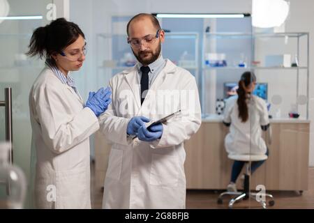 Chemist nurse explainig to doctor vaccine development in modern equipped laboratory pointing on tablet. Team of doctors examining virus evolution using high tech researching diagnosis. Stock Photo