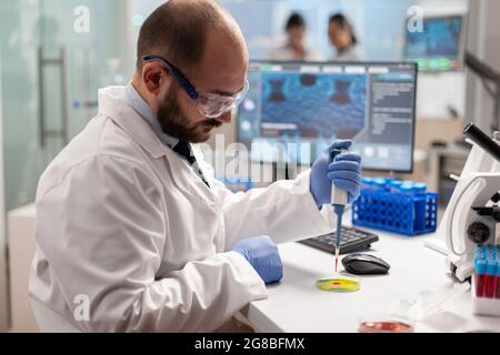 Scientist dropping blood sample with micropipette on petri dish in genetic laboratory. Team of researchers examining virus evolution using high tech for vaccine development. Stock Photo