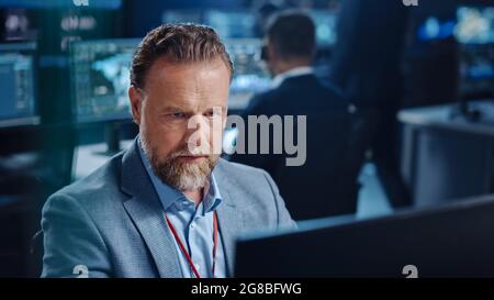 Portrait of Serious Professional Technical Controller Sitting at His Desk with Multiple Computer Displays Before Him. In the Background His Colleagues Stock Photo