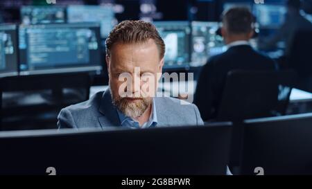 Portrait of Positive Professional Technical Controller Sitting at His Desk with Multiple Computer Displays Before Him. In the Background His Stock Photo