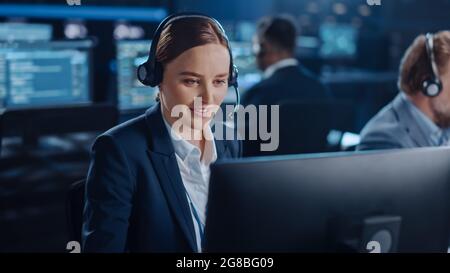 Joyful Beautiful Technical Customer Support Specialist is Talking on a Headset while Working on a Computer in a Dark Monitoring and Control Room Stock Photo