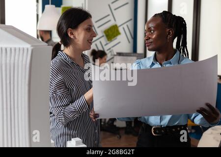 Colleagues architects women checking plans of blueprints layout design. Multi ethnic team of professional workers standing at building model maquette for modern technology project Stock Photo