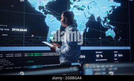 Project Manager Walks to a Big Screen with Global Map. He is Using a Tablet. Team of Professional Computer Data Science Engineers Work in a Dark Stock Photo