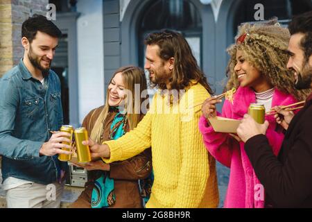 Best friends gathering in the street eating take away food with noodles and toasting with canned drinks such as beers Stock Photo