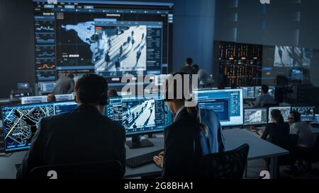 Female Officer Works on a Computer with Surveillance CCTV Video Footage in a Police Monitoring Center with Global Map Tracking on a Big Digital Screen Stock Photo