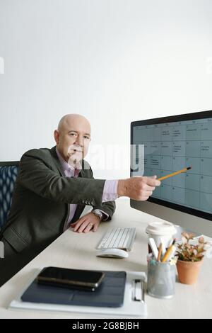 Portrait of mature businessman looking at camera while sitting at his workplace and pointing at computer monitor Stock Photo